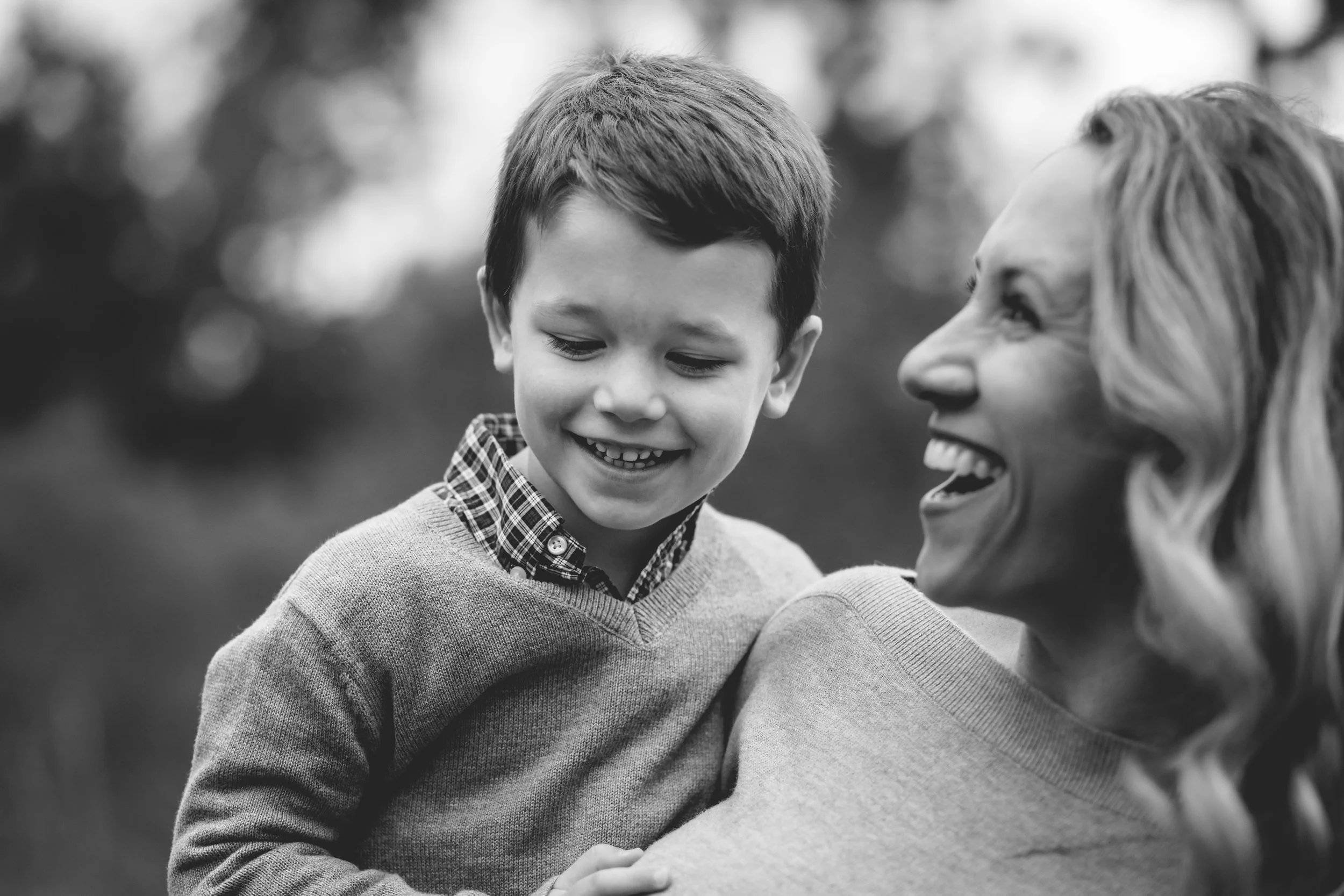 A woman and a young boy smiling and looking at each other outdoors in a black and white photograph.