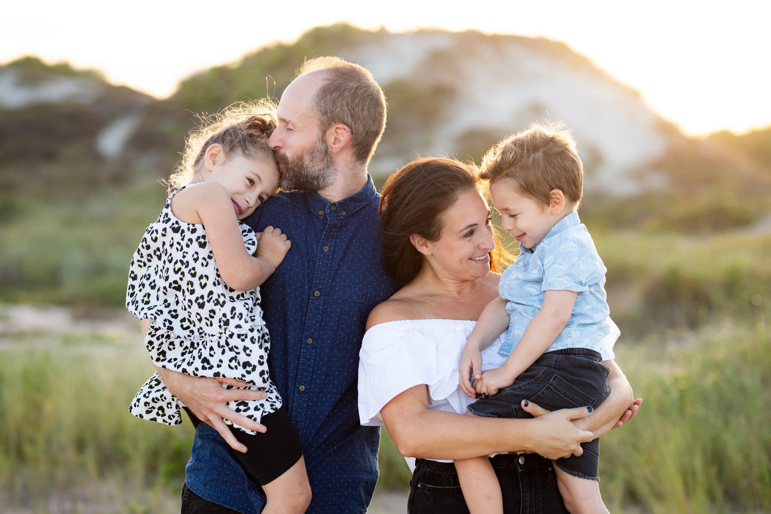 A family of four outdoors during sunset, with parents each holding a child and sharing a tender moment, surrounded by green grass and hills.