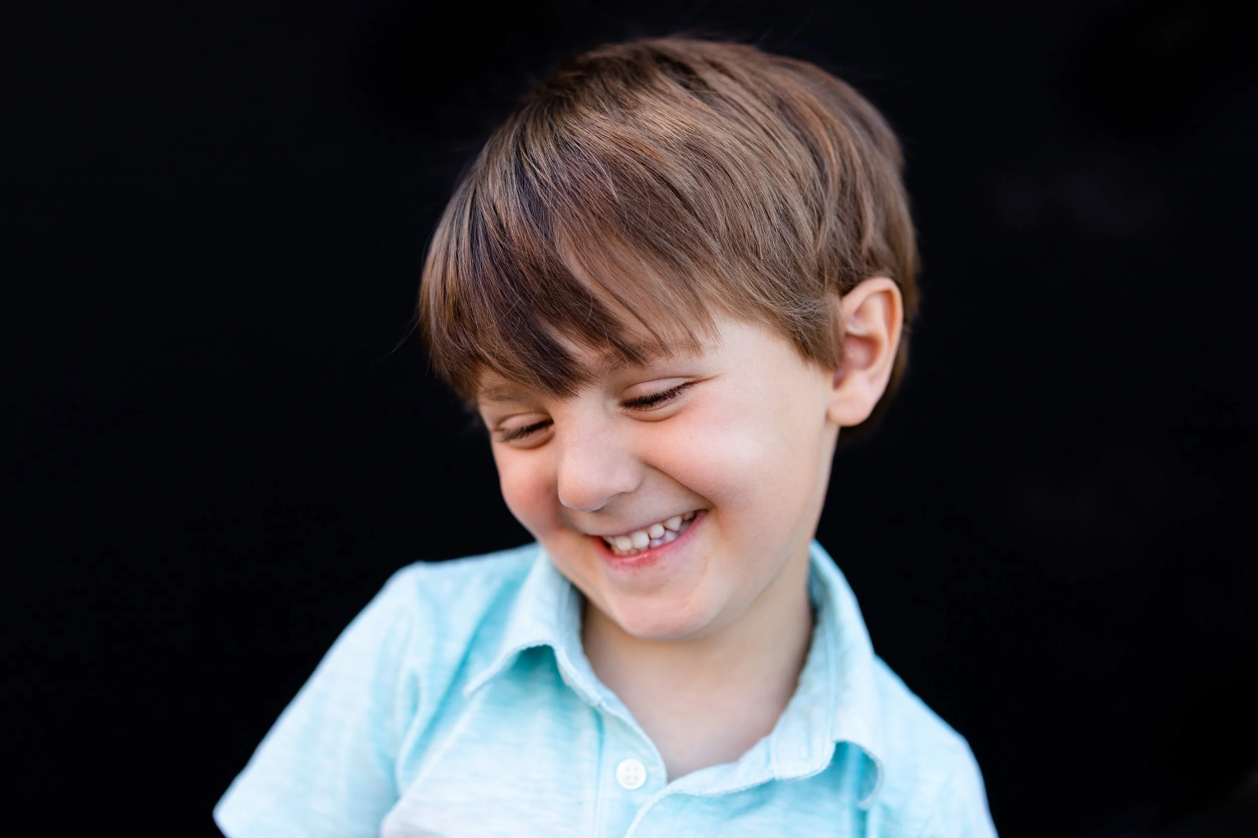 A young boy with brown hair smiling and looking down, wearing a light blue shirt, against a black background.
