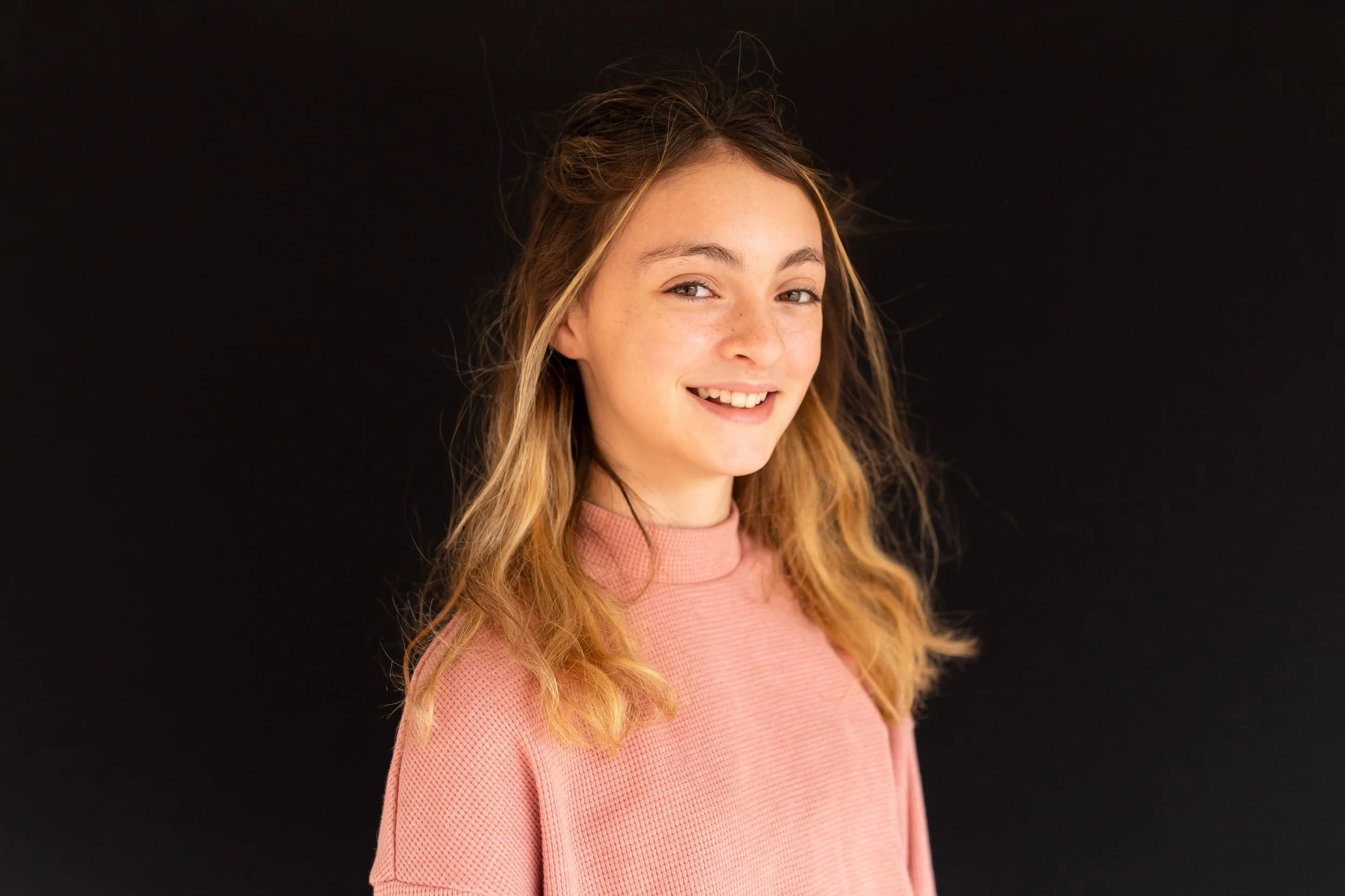Portrait of a young woman with wavy blonde hair, smiling, wearing a pink textured top, against a black background.