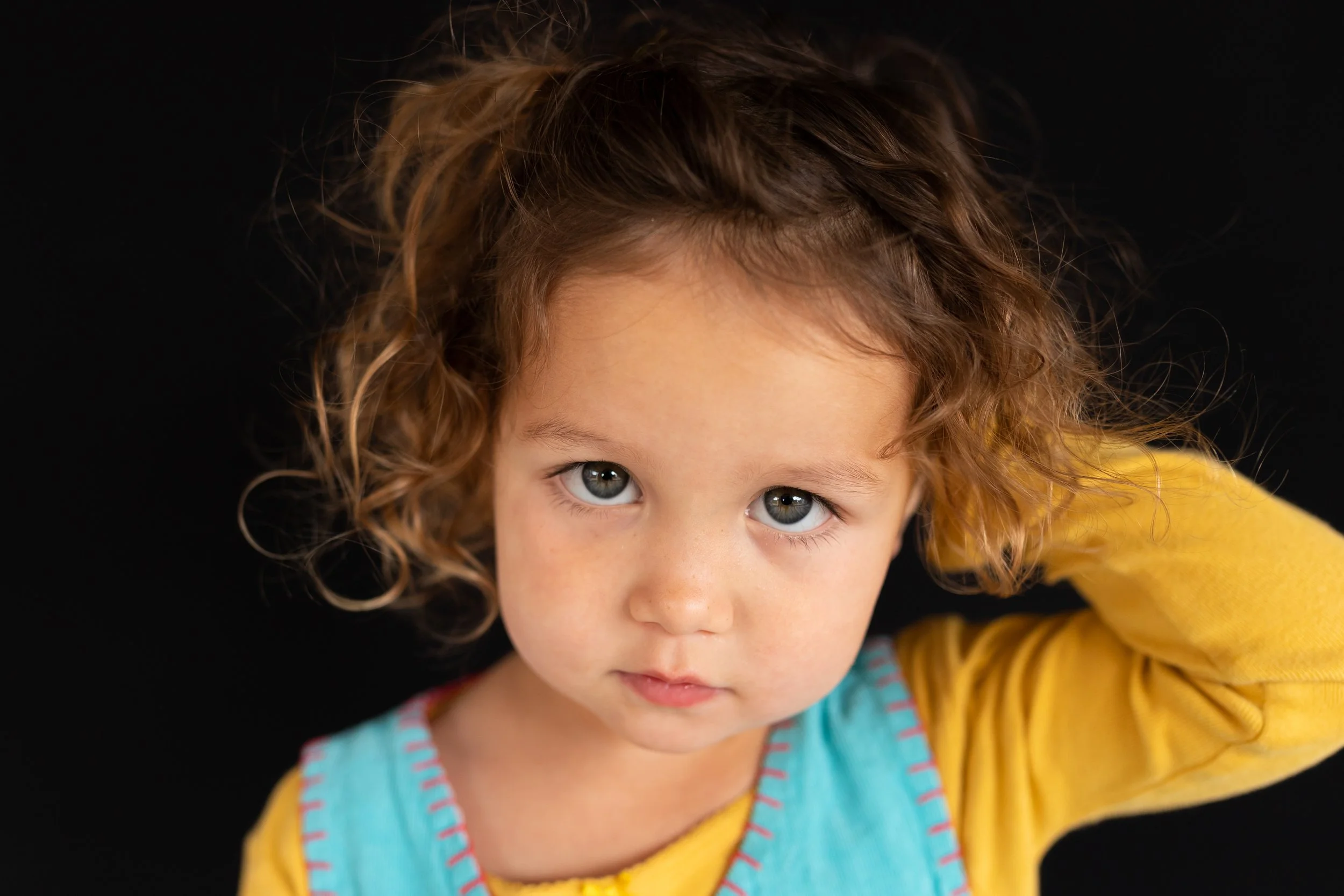 A young girl with curly light brown hair and blue eyes wearing a yellow shirt and a blue bib, looking into the camera with a serious expression, against a black background.