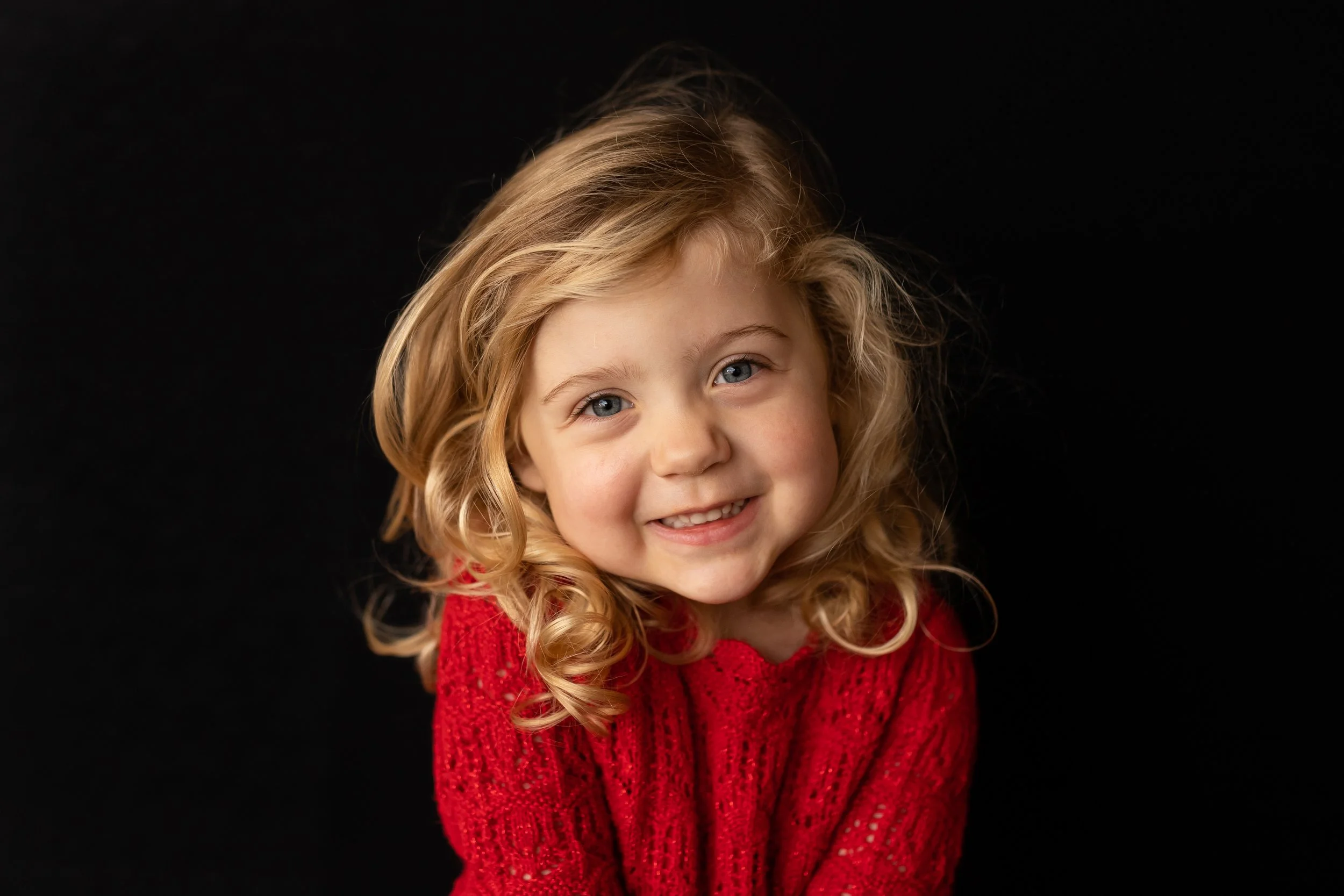 A young girl with blonde curly hair and blue eyes, smiling and wearing a red sweater, against a black background.