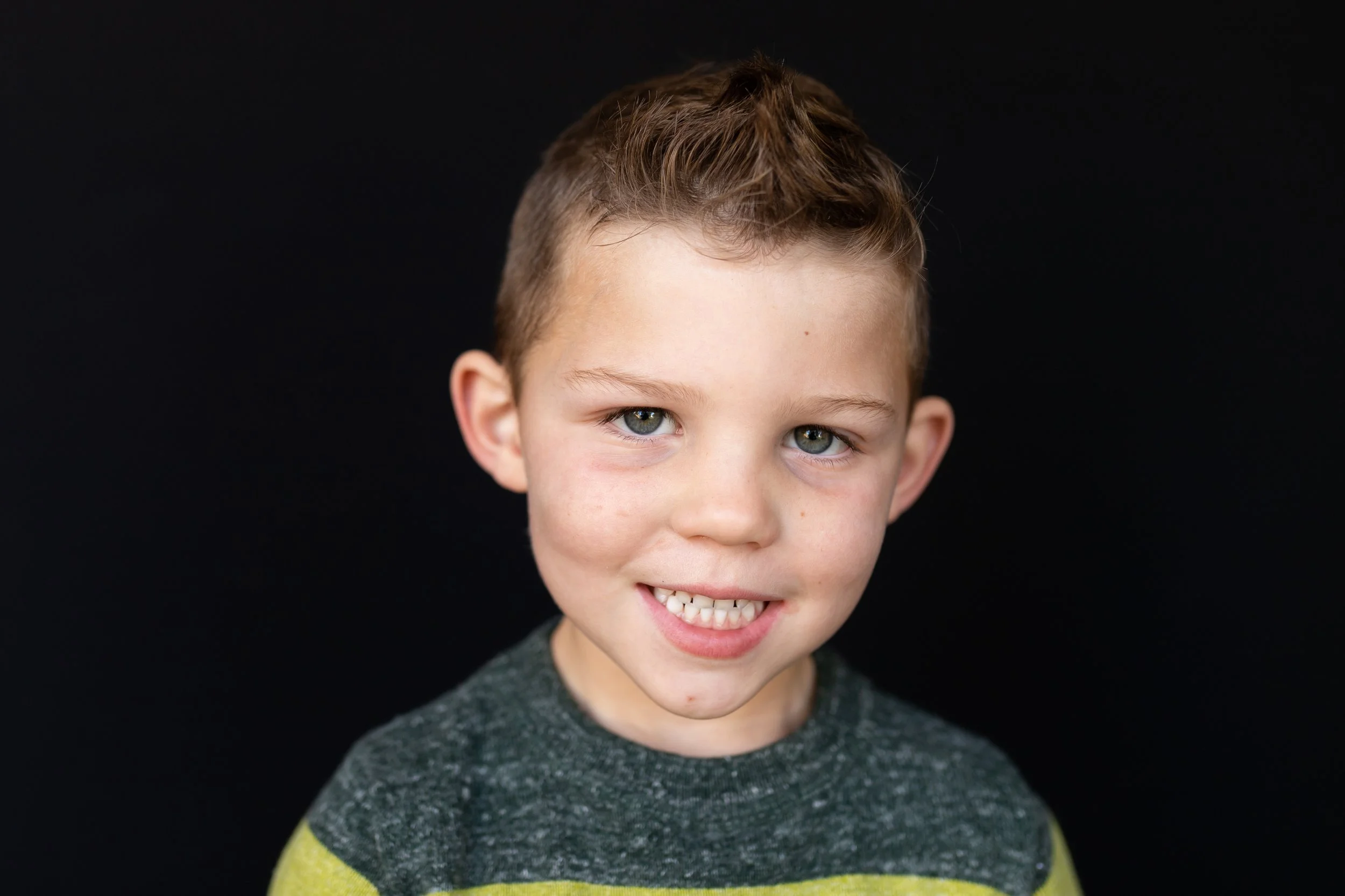 A young boy with short light brown hair, blue eyes, and a slight smile, wearing a dark gray and yellow shirt, standing against a black background.