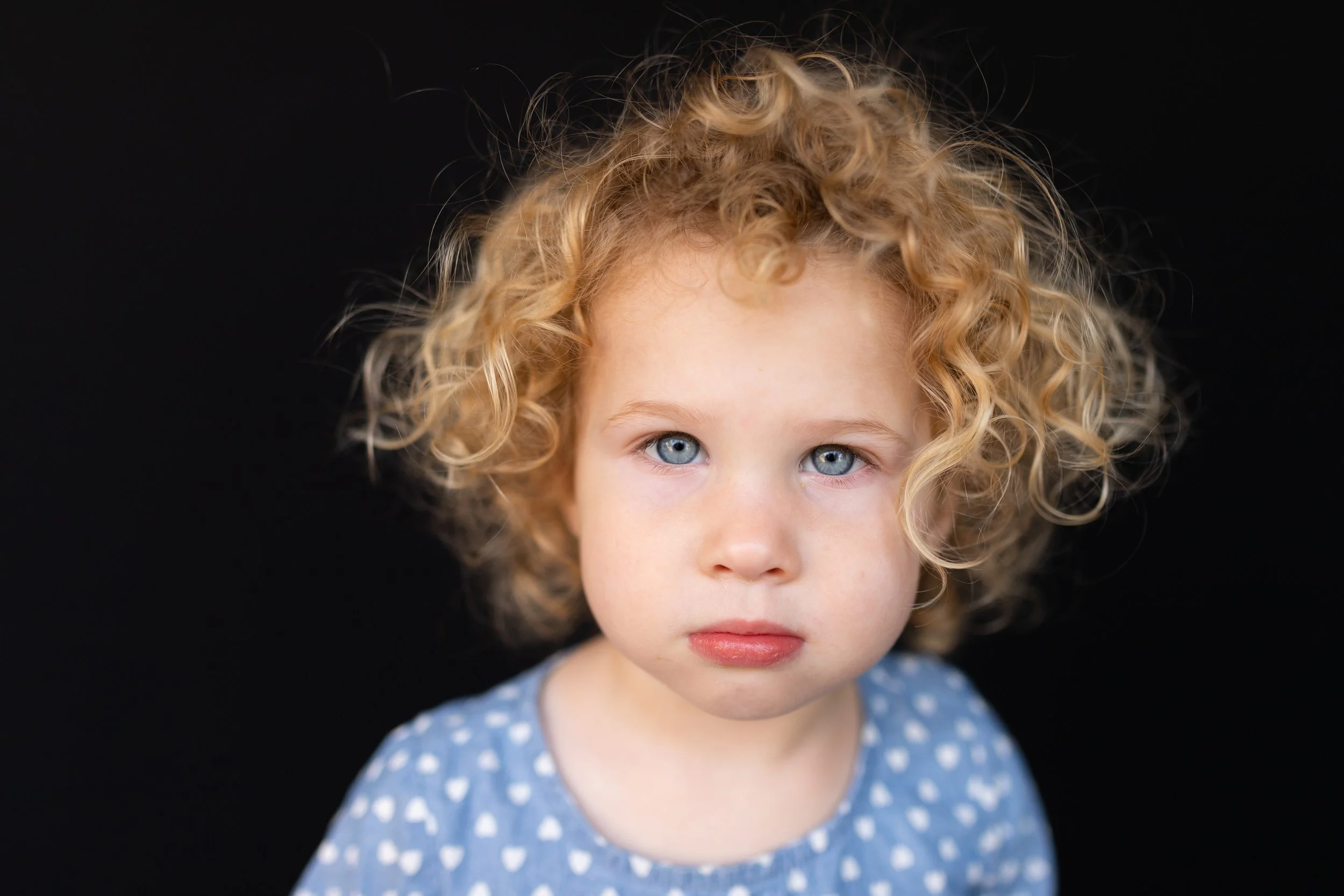 A young girl with curly blonde hair and blue eyes, wearing a blue dress with white polka dots, looking directly at the camera with a neutral expression, against a black background.