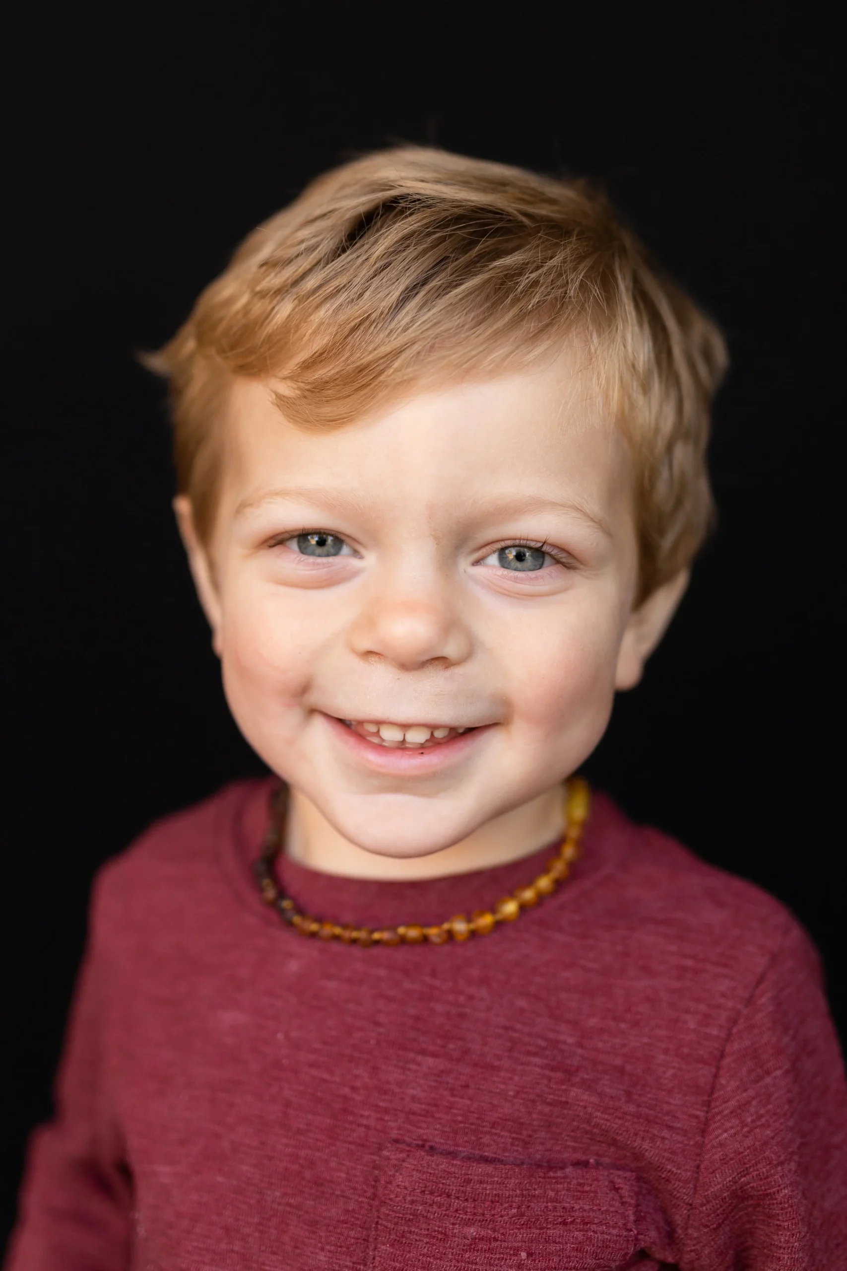 Close-up portrait of a young boy with red hair, blue eyes, smiling, wearing a maroon shirt and a yellow beaded necklace, against a black background.