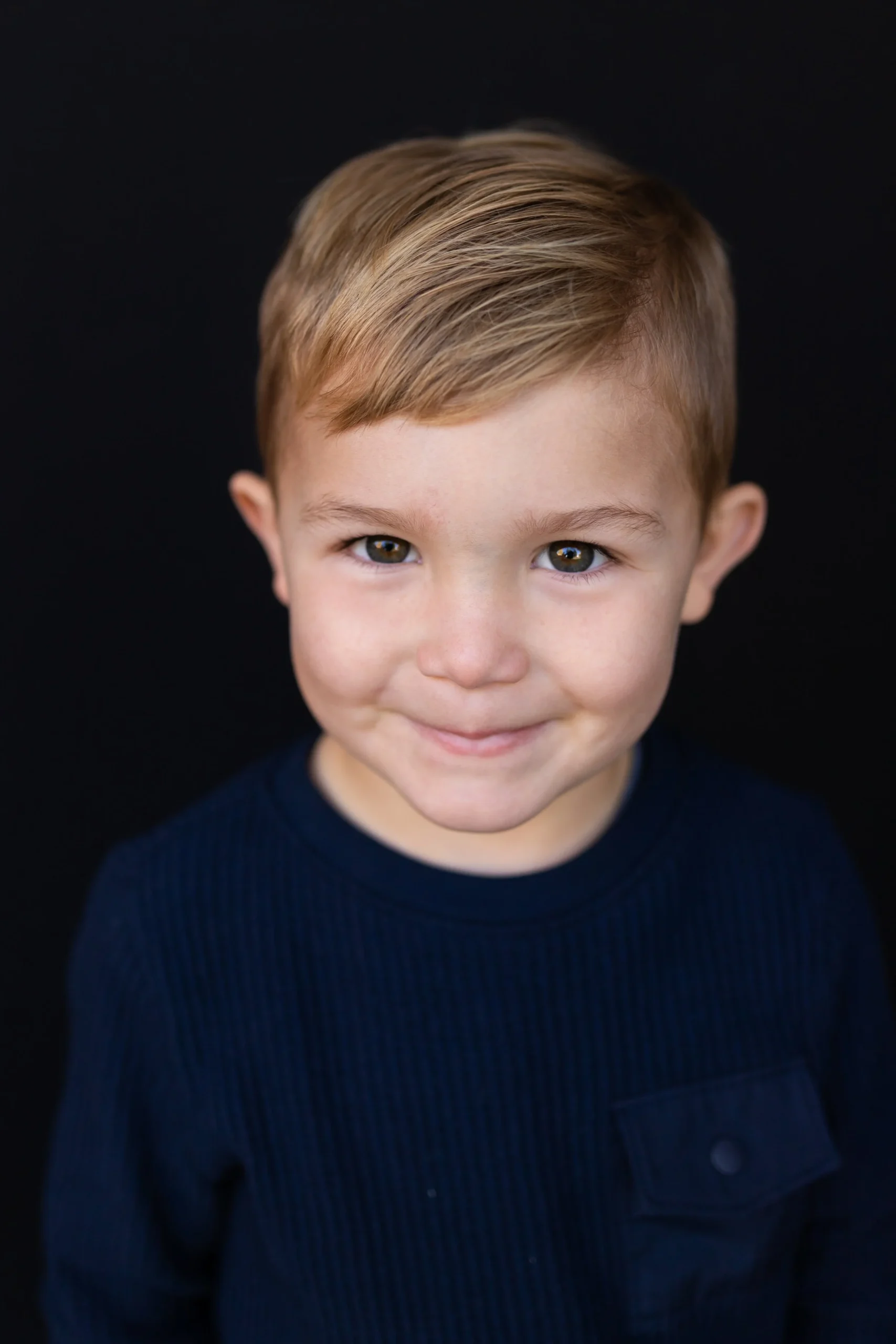 Close-up of a young boy with light brown hair and hazel eyes, smiling slightly, wearing a navy blue shirt, against a black background.