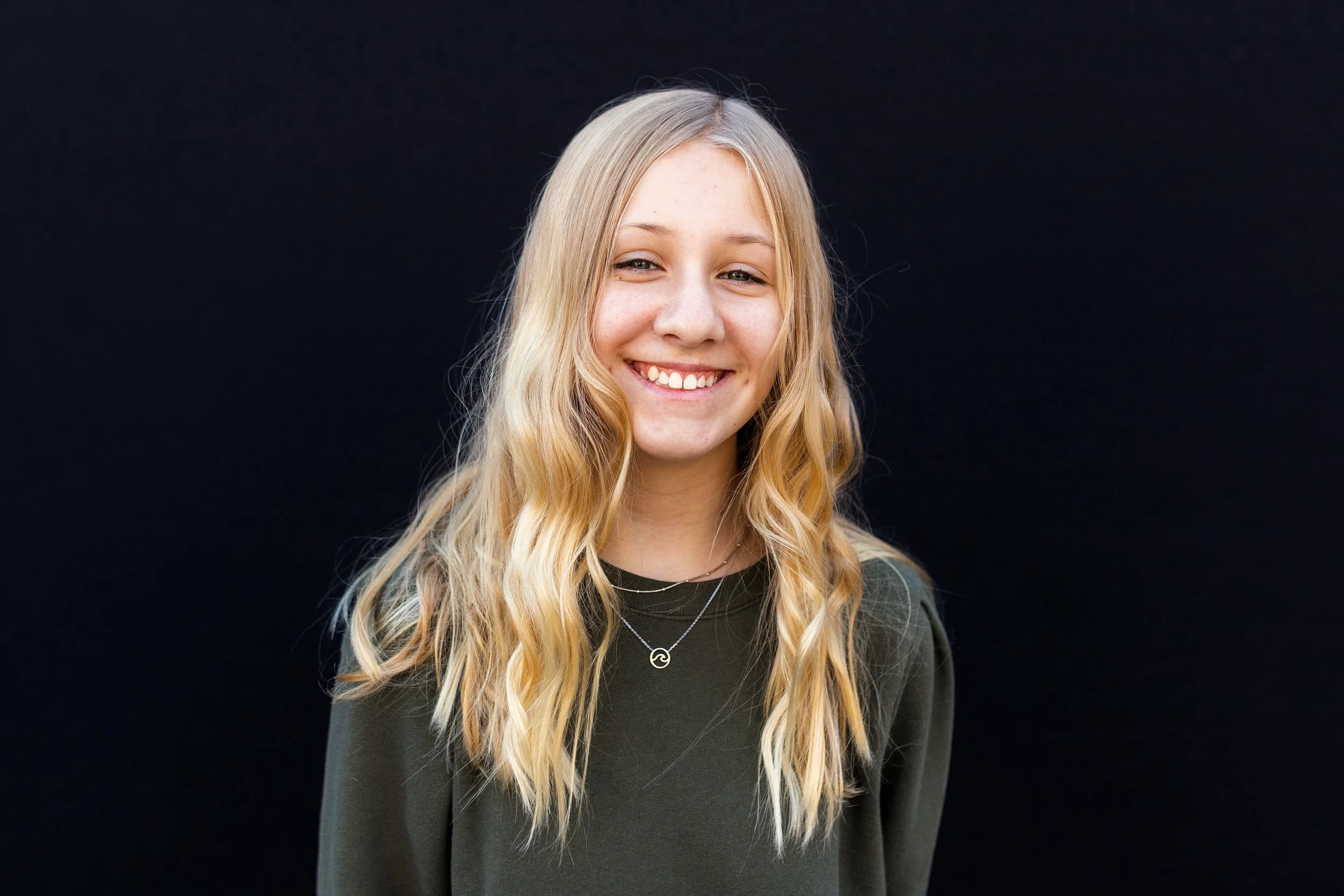 Smiling young woman with blonde wavy hair standing against a black background.