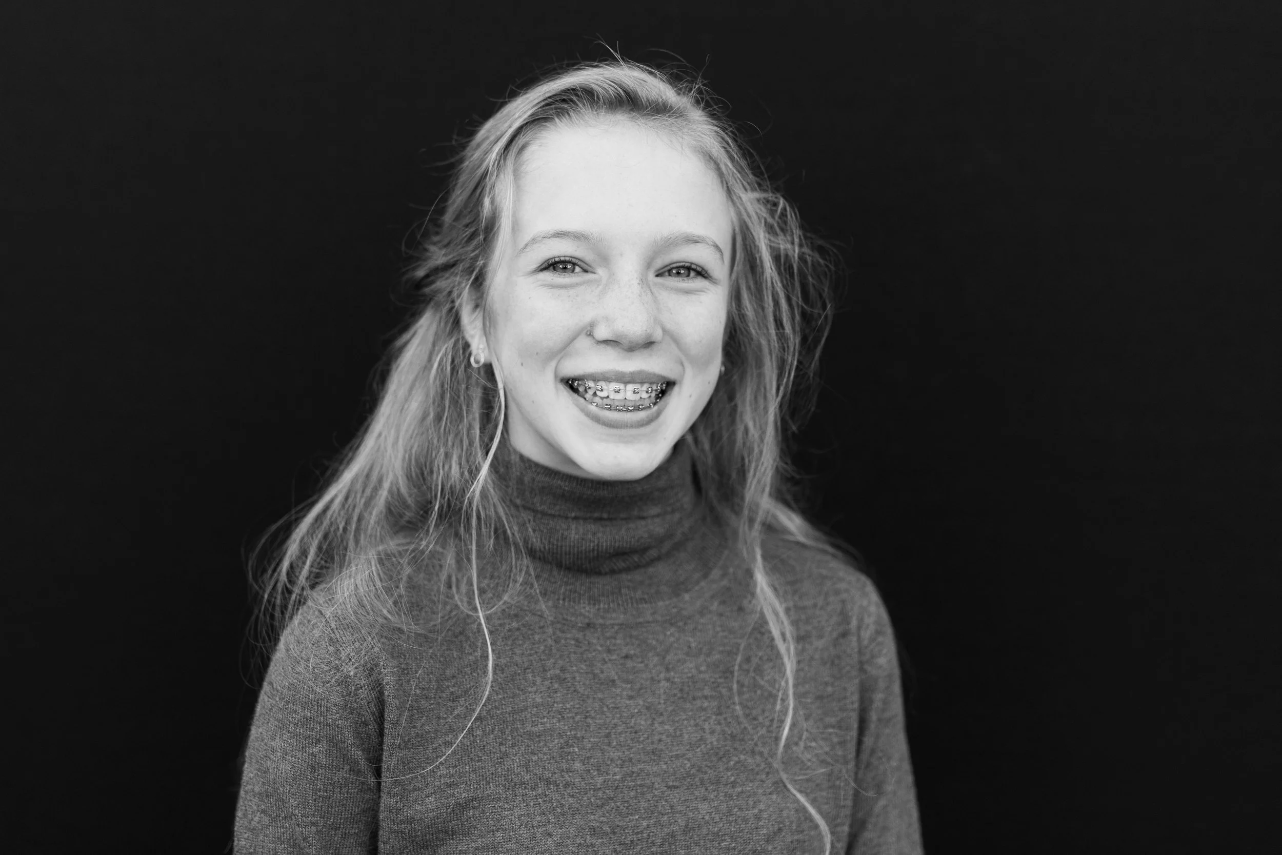 Black and white photo of a young woman with long, wavy hair smiling with braces, wearing a turtleneck sweater, against a dark background.