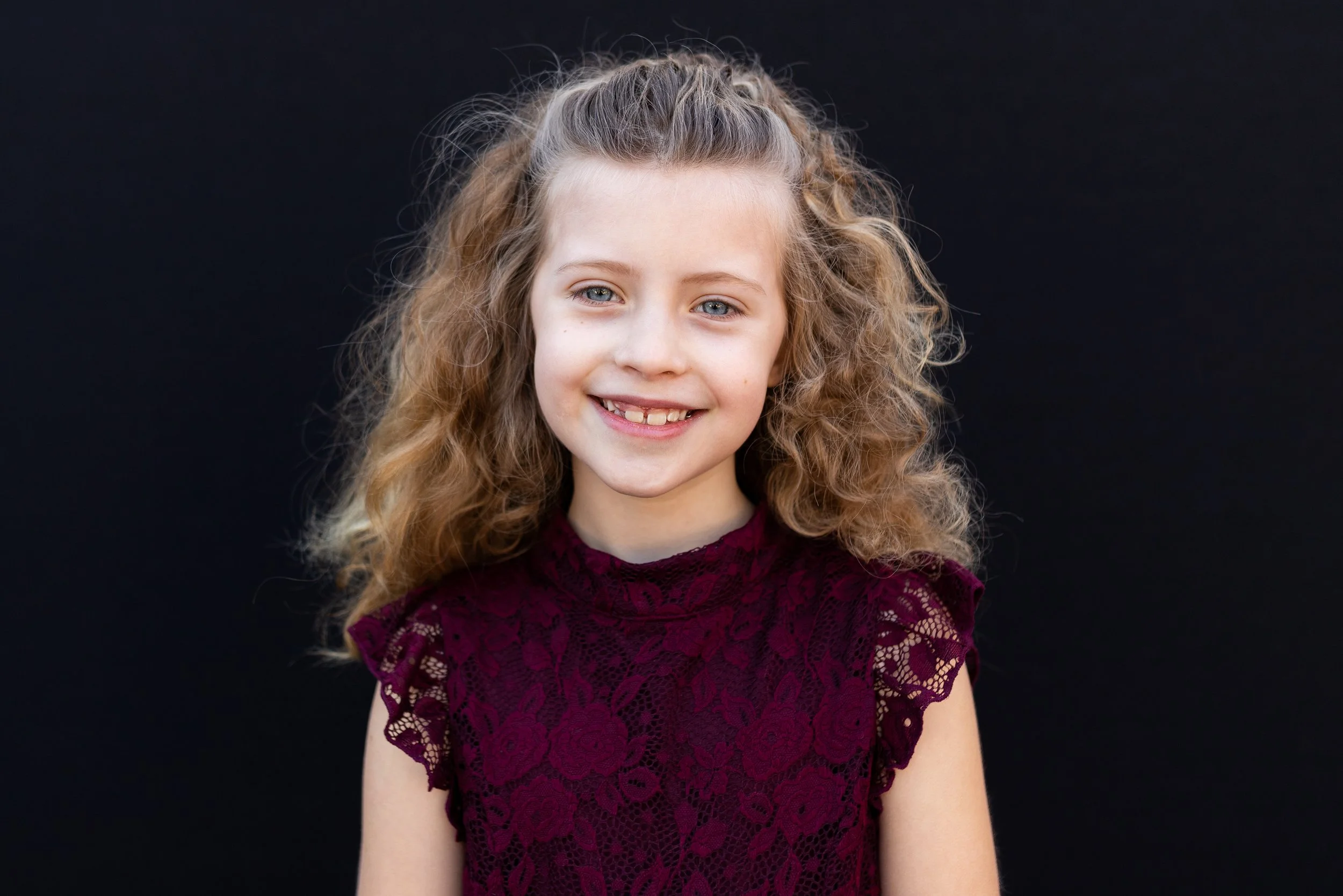 A young girl with curly blonde hair and blue eyes wearing a maroon lace top, smiling at the camera against a black background.