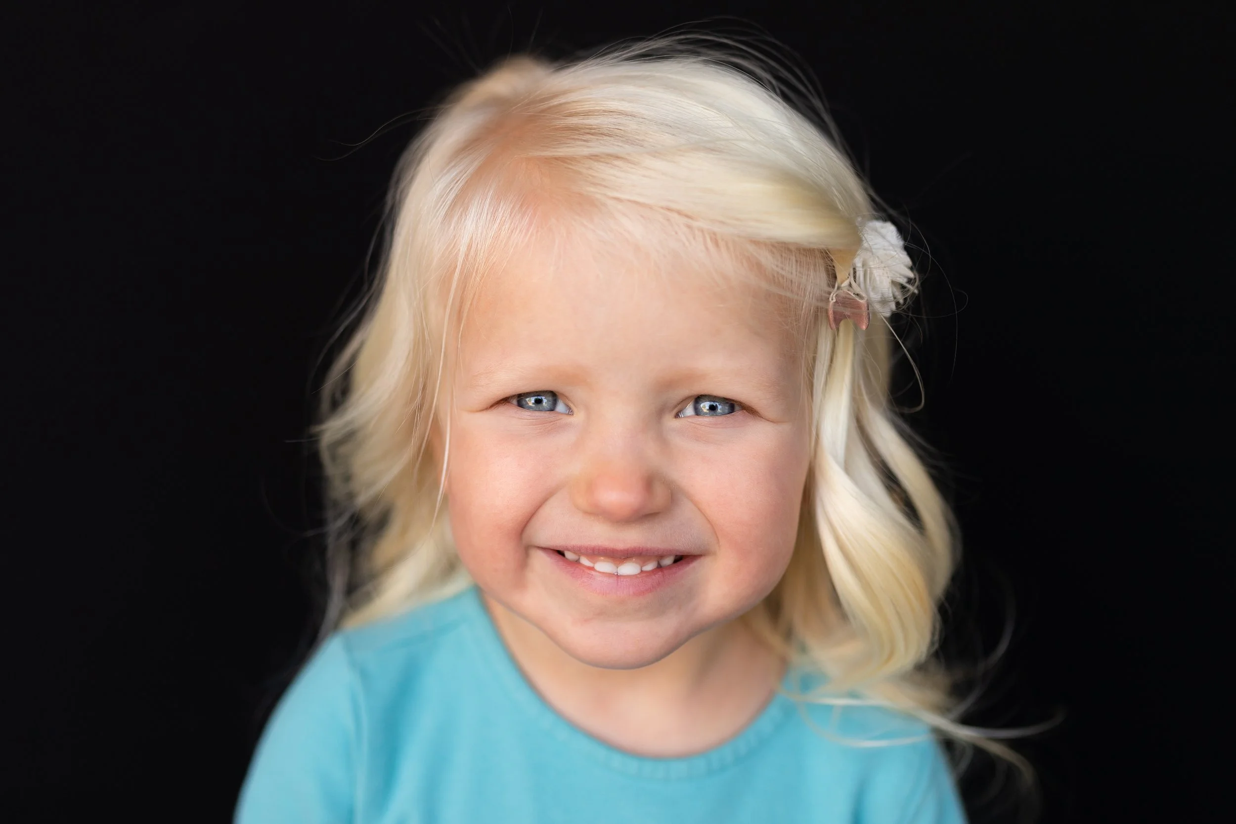 A close-up portrait of a young girl with blonde hair, blue eyes, smiling, wearing a turquoise shirt, against a black background.